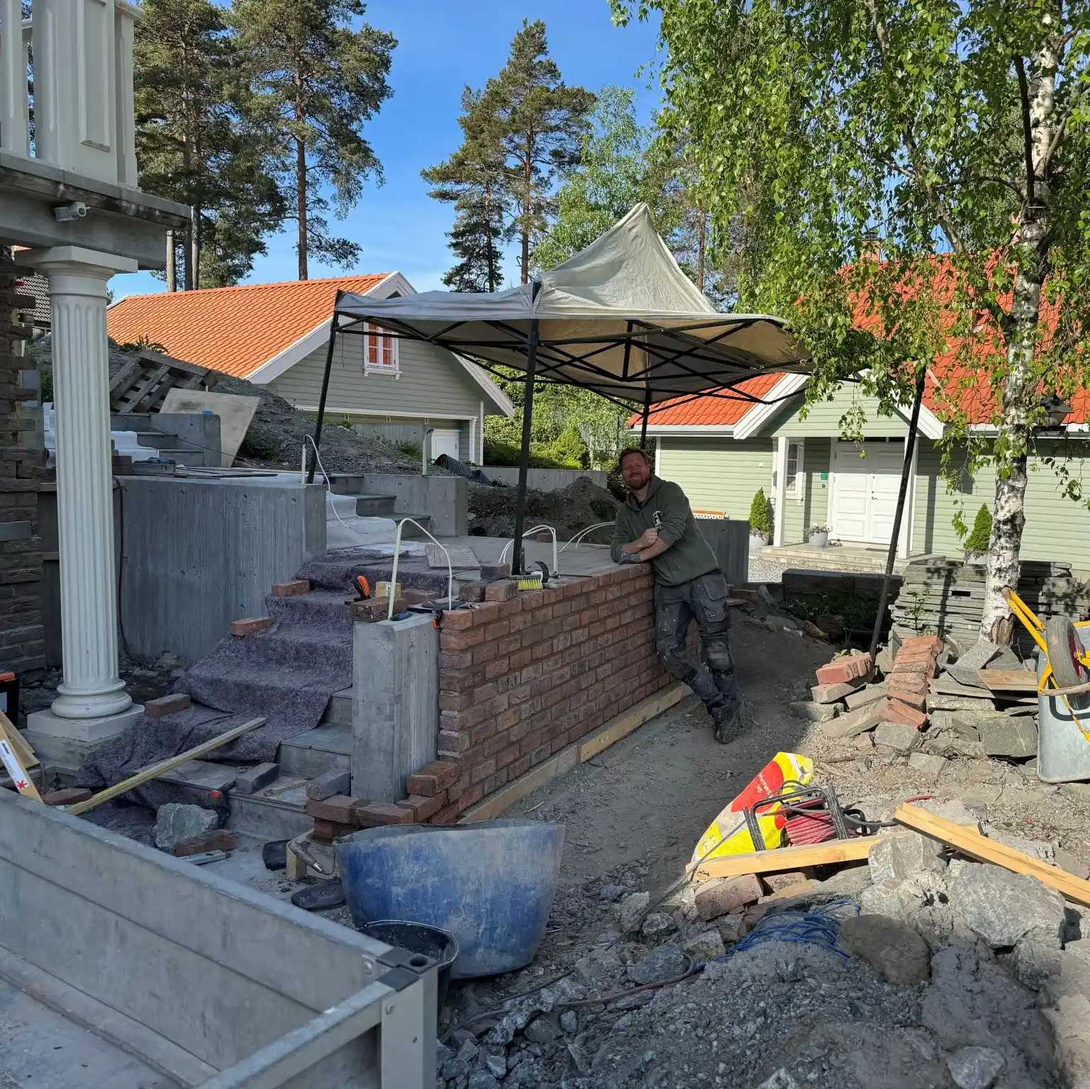 En mann lener seg mot en murvegg under bygging ved siden av en steintrapp. Verktøy og materialer ligger spredt rundt, og huset i bakgrunnen har grønn fasade og rødt tak.