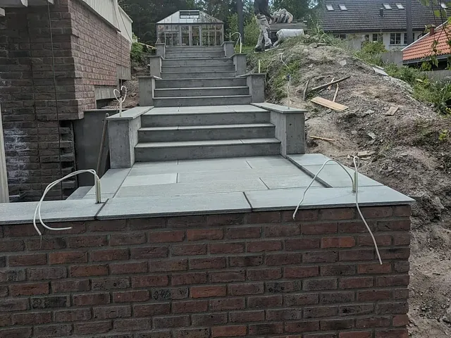 En bred steintrapp med åtte trinn fører opp til et drivhus. En mann arbeider på toppen av trappen, omgitt av trær og byggeutstyr.