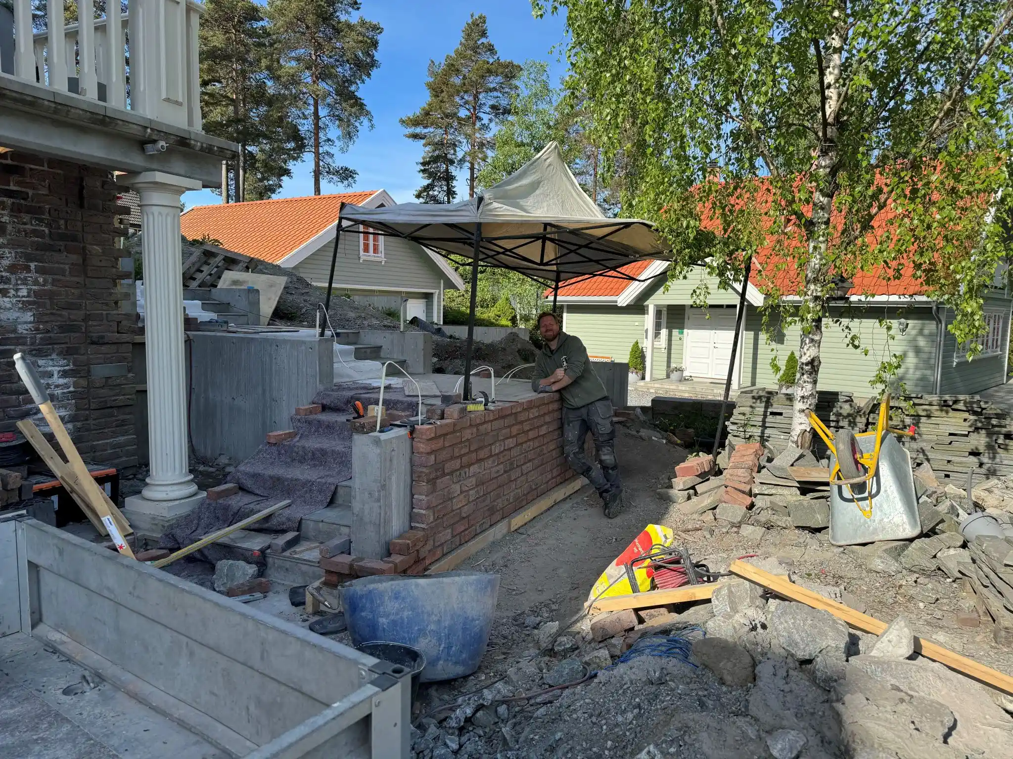 En mann lener seg mot en murvegg under bygging ved siden av en steintrapp. Verktøy og materialer ligger spredt rundt, og huset i bakgrunnen har grønn fasade og rødt tak.