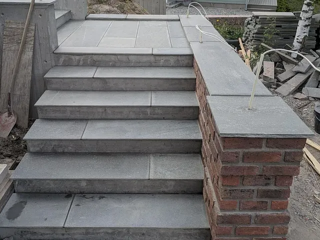 En steintrapp med seks trinn leder opp til et hus med grå fasade og rødt tak. Trappen har en murvegg med teglstein på høyre side og er omringet av en byggeplass.