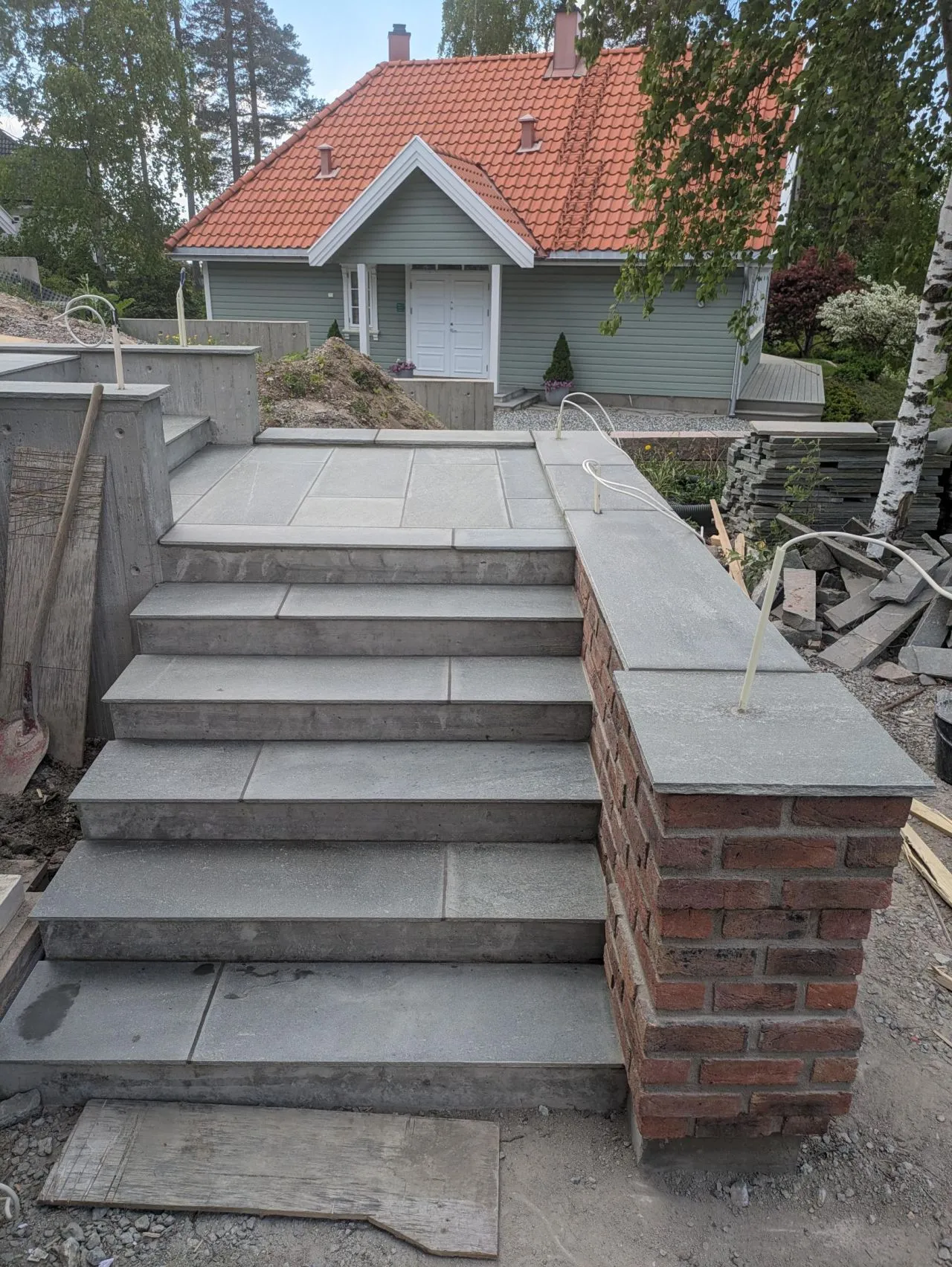 En steintrapp med seks trinn leder opp til et hus med grå fasade og rødt tak. Trappen har en murvegg med teglstein på høyre side og er omringet av en byggeplass.