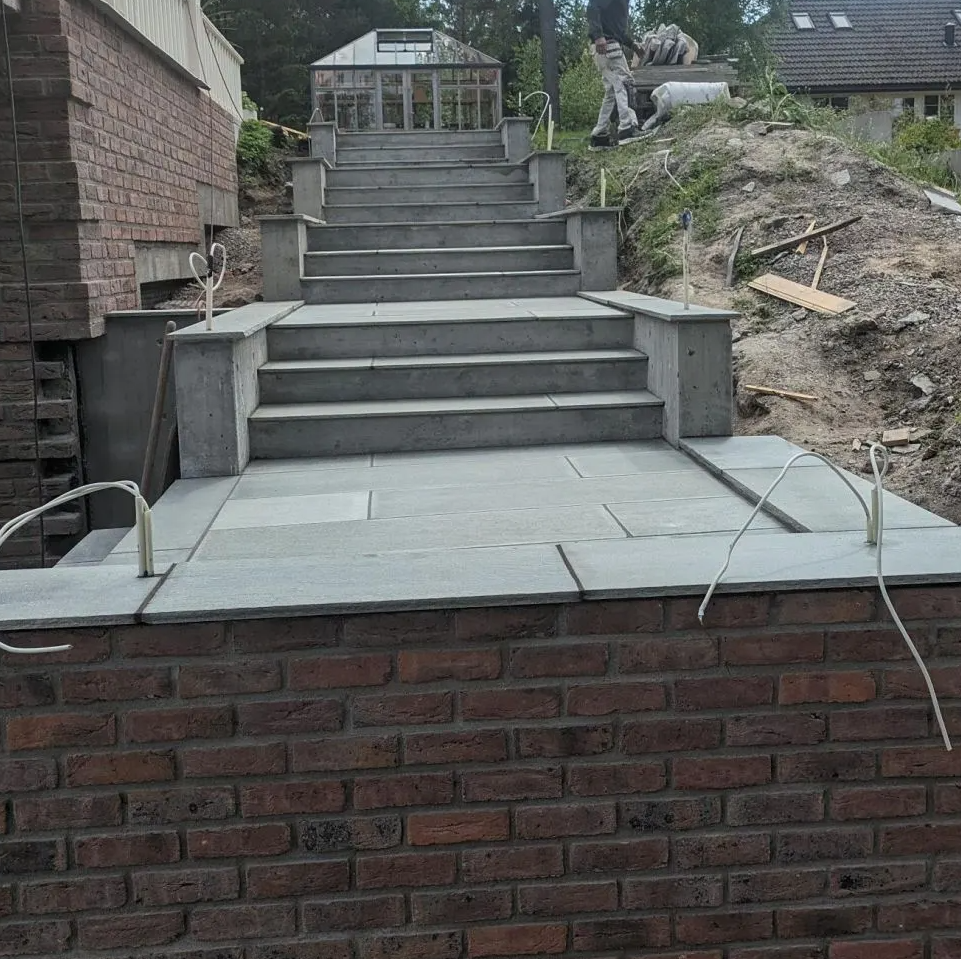 En bred steintrapp med åtte trinn fører opp til et drivhus. En mann arbeider på toppen av trappen, omgitt av trær og byggeutstyr.