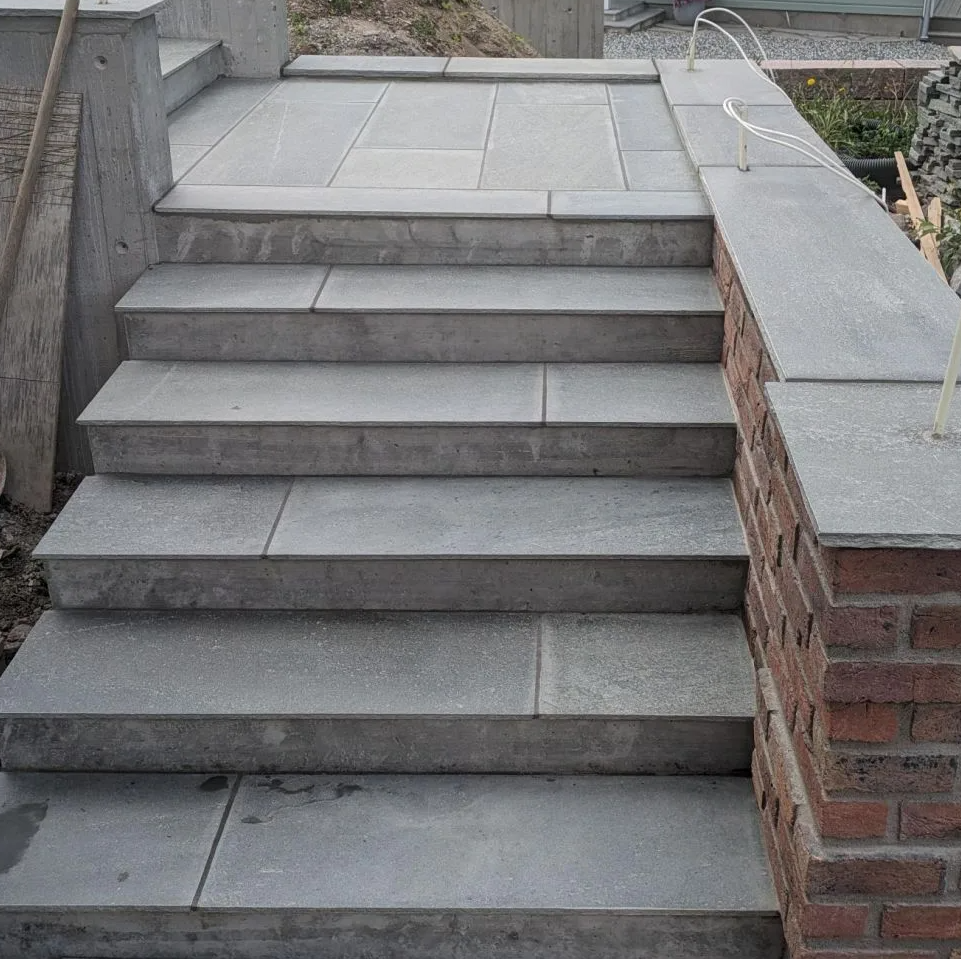 En steintrapp med seks trinn leder opp til et hus med grå fasade og rødt tak. Trappen har en murvegg med teglstein på høyre side og er omringet av en byggeplass.