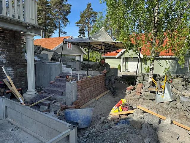 En mann lener seg mot en murvegg under bygging ved siden av en steintrapp. Verktøy og materialer ligger spredt rundt, og huset i bakgrunnen har grønn fasade og rødt tak.