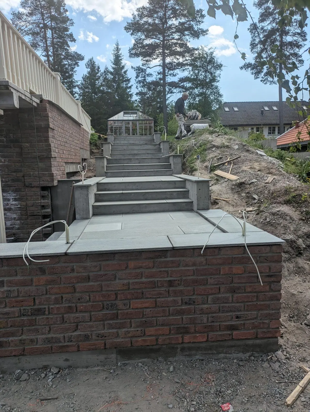 En bred steintrapp med åtte trinn fører opp til et drivhus. En mann arbeider på toppen av trappen, omgitt av trær og byggeutstyr.