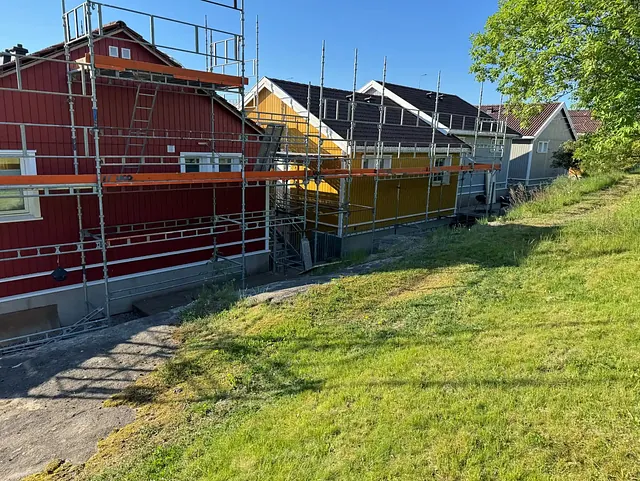Rød trebygning med stillas langs veggen, under renovering, med gult hus i bakgrunnen og en stor grønn plen på høyre side.