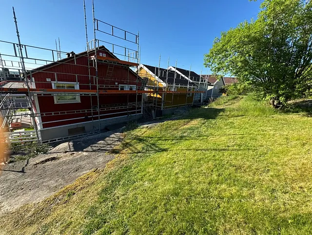 Bredere bilde av den røde bygningen med stillas, viser mer av den grønne plenen og et stort tre til høyre under en klar blå himmel.